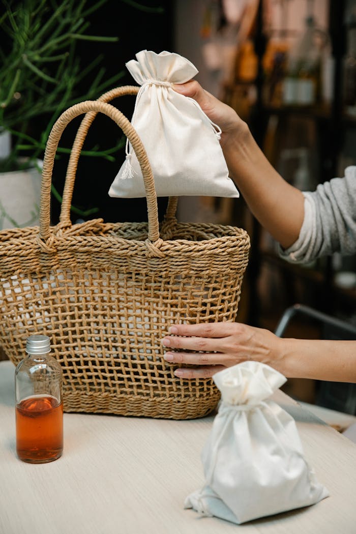 gallery-5 Hands placing a fabric pouch into a woven basket with a glass bottle nearby on a wooden table.