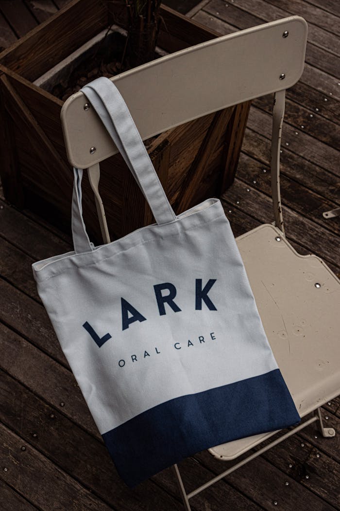 gallery-2 A modern tote bag with 'Lark Oral Care' branding hangs on a white chair in an outdoor setting.